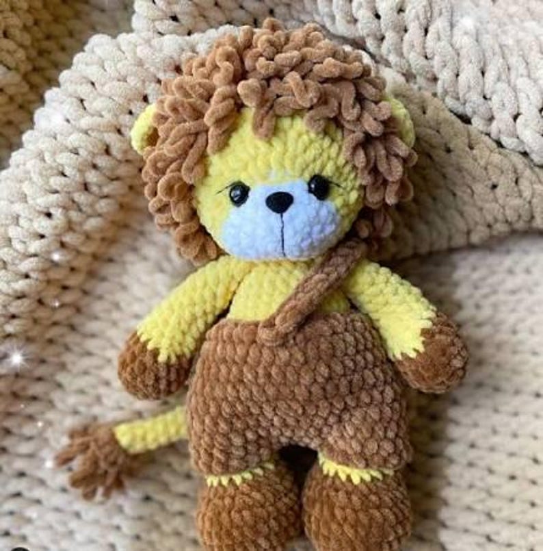 May include: A handmade crochet lion stuffed animal. The lion has a yellow body, brown mane, and brown overalls. The face is yellow and blue with black eyes and a black nose. The lion is sitting on a cream-colored blanket.