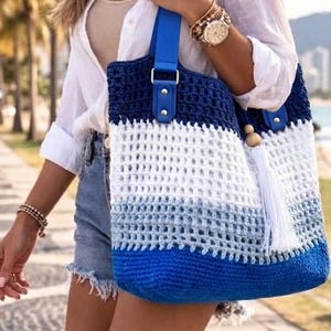 May include: A crochet tote bag featuring a horizontal color-blocked design in dark blue, white, and light blue. The bag has a bright blue adjustable shoulder strap attached with silver-toned hardware and a white tassel with a wooden bead accent.