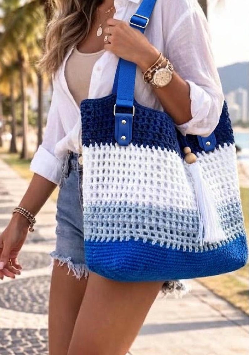 May include: A crochet tote bag featuring a horizontal color-blocked design in dark blue, white, and light blue. The bag has a bright blue adjustable shoulder strap attached with silver-toned hardware and a white tassel with a wooden bead accent.
