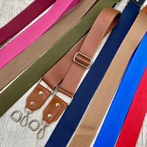 May include: A collection of adjustable bag straps in various solid colors including burgundy, pink, olive green, tan, navy blue, bright blue, and red. Each strap features a woven fabric texture, silver-toned metal hardware, and brown leather tabs.