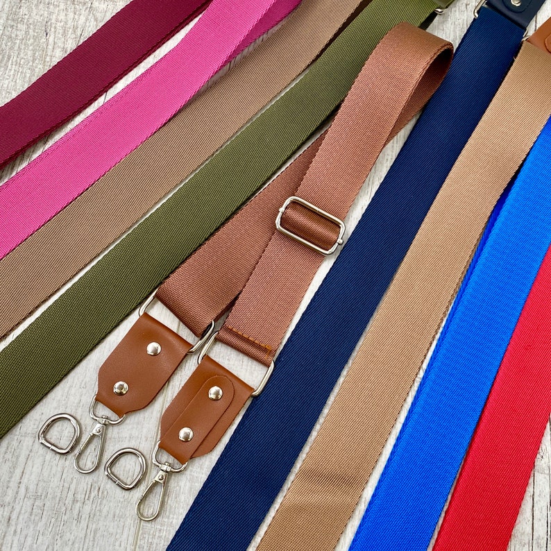 May include: A collection of adjustable bag straps in various solid colors including burgundy, pink, olive green, tan, navy blue, bright blue, and red. Each strap features a woven fabric texture, silver-toned metal hardware, and brown leather tabs.