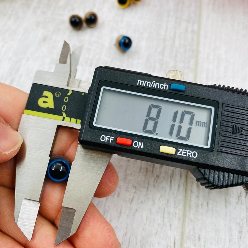 May include: A small, round, blue and black plastic safety eye being measured by a digital caliper. The digital display on the black caliper clearly shows a measurement of 8.10 mm. Several other similar safety eyes are visible in the background.