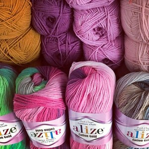 May include: A collection of Alize Cotton Gold yarn skeins in various colors including mustard yellow, purple, pink, and variegated blends. Each skein features a paper label with the brand name Alize Cotton Gold printed in black and purple text.