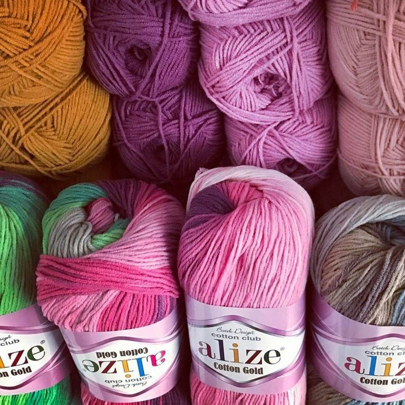 May include: A collection of Alize Cotton Gold yarn skeins in various colors including mustard yellow, purple, pink, and variegated blends. Each skein features a paper label with the brand name Alize Cotton Gold printed in black and purple text.