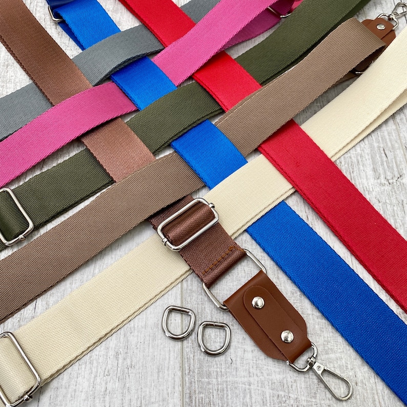 May include: A collection of adjustable bag straps in various colors including red, blue, olive green, brown, pink, gray, and cream. Each strap features silver-toned metal hardware, including sliders and swivel lobster clasps, with some straps featuring brown faux leather accents.