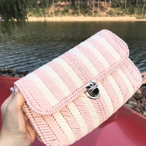 May include: A pink and white striped handbag with a silver clasp. The bag has a woven texture and a rectangular shape. The background shows a lake and trees.