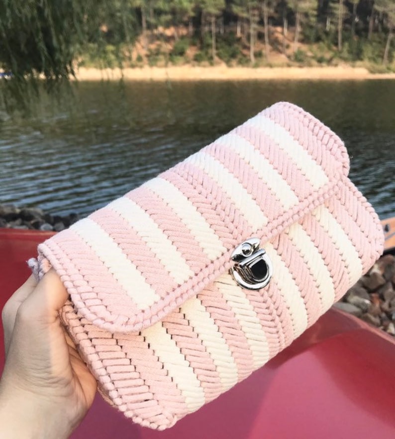 May include: A pink and white striped handbag with a silver clasp. The bag has a woven texture and a rectangular shape. The background shows a lake and trees.