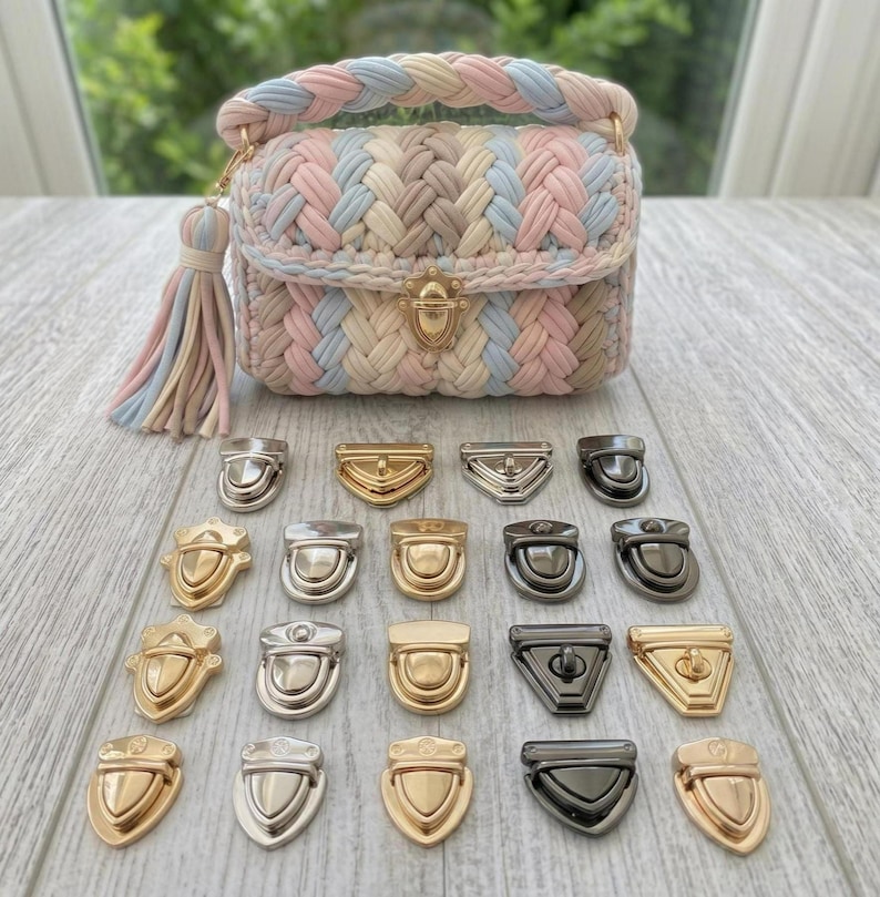May include: A pastel-coloured crochet handbag with a braided handle and tassel, featuring a gold-tone clasp. Assorted metal clasps in gold, silver, and black are displayed below the bag, showcasing various shapes and designs. The bag is approximately 18 cm wide.