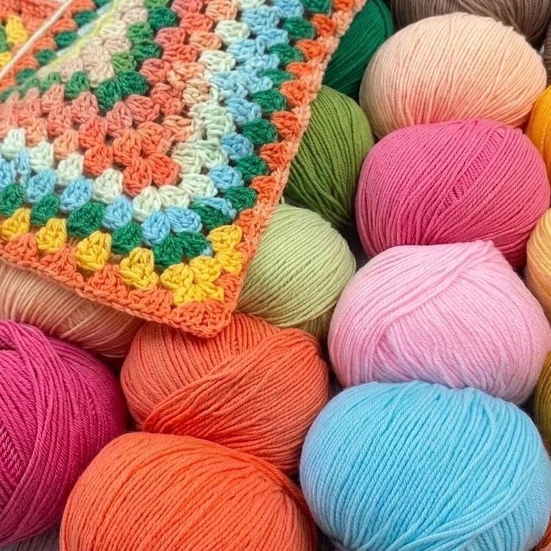 May include: A collection of multi-colored yarn balls in shades of pink, orange, yellow, green, and blue, arranged alongside a partially completed crochet square featuring a granny square pattern in matching vibrant yarn colors.