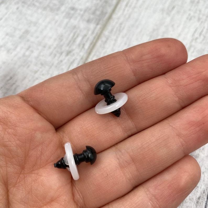May include: Two black plastic safety eyes with smooth, rounded tops and threaded stems, each paired with a translucent white plastic washer. These small components are designed for securing onto fabric or plush surfaces for craft projects.