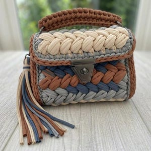 May include: A handmade crochet handbag in shades of brown, beige, grey, and blue. The bag features a braided design, a top handle, and a metal clasp. A tassel of matching colours hangs from the side. The bag is a stylish accessory.