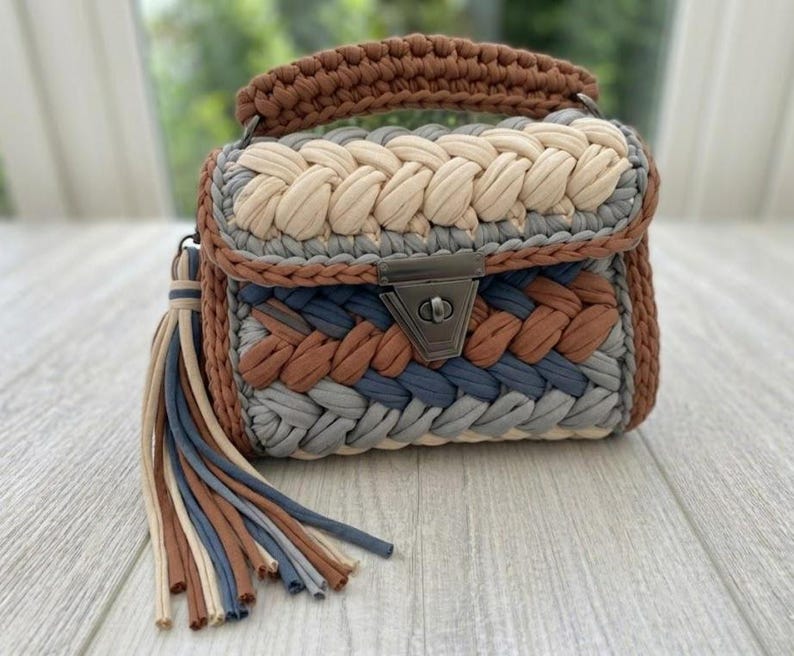 May include: A handmade crochet handbag in shades of brown, beige, grey, and blue. The bag features a braided design, a top handle, and a metal clasp. A tassel of matching colours hangs from the side. The bag is a stylish accessory.