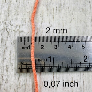 Może przedstawiać: Zbliżenie na pomarańczową nić o średnicy 2 mm, obok metalowej linijki. Linijka pokazuje zar&oacute;wno pomiary w calach, jak i w centymetrach. Widoczny tekst "2 mm" i "0,07 inch".