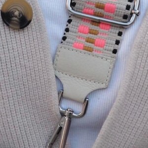 May include: A woven bag strap featuring a pattern of neon pink, tan, and black rectangular blocks on a light beige background. The strap includes a silver metal adjustable slider, a light beige leather tab, and a silver metal lobster clasp.