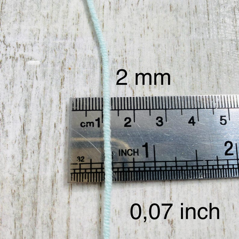 May include: A single strand of light mint green cord positioned vertically against a metal ruler. The ruler displays measurements in both centimeters and inches, clearly indicating the cord has a diameter of 2 mm or 0.07 inches.
