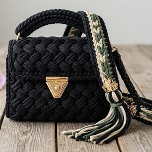 May include: A black crochet handbag with a woven pattern. The bag features a gold-coloured clasp and a braided handle. The shoulder strap is a mix of black, green, and beige. A tassel hangs from the strap.