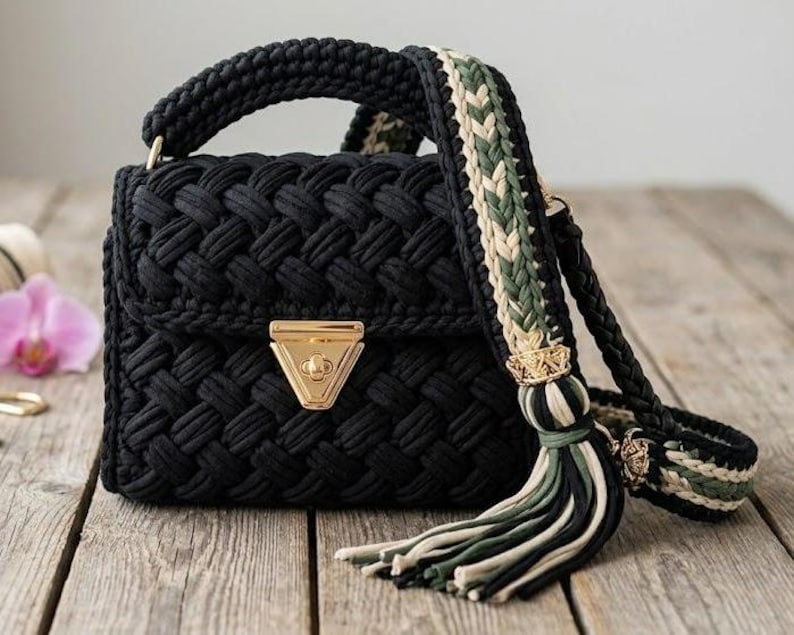 May include: A black crochet handbag with a woven pattern. The bag features a gold-coloured clasp and a braided handle. The shoulder strap is a mix of black, green, and beige. A tassel hangs from the strap.