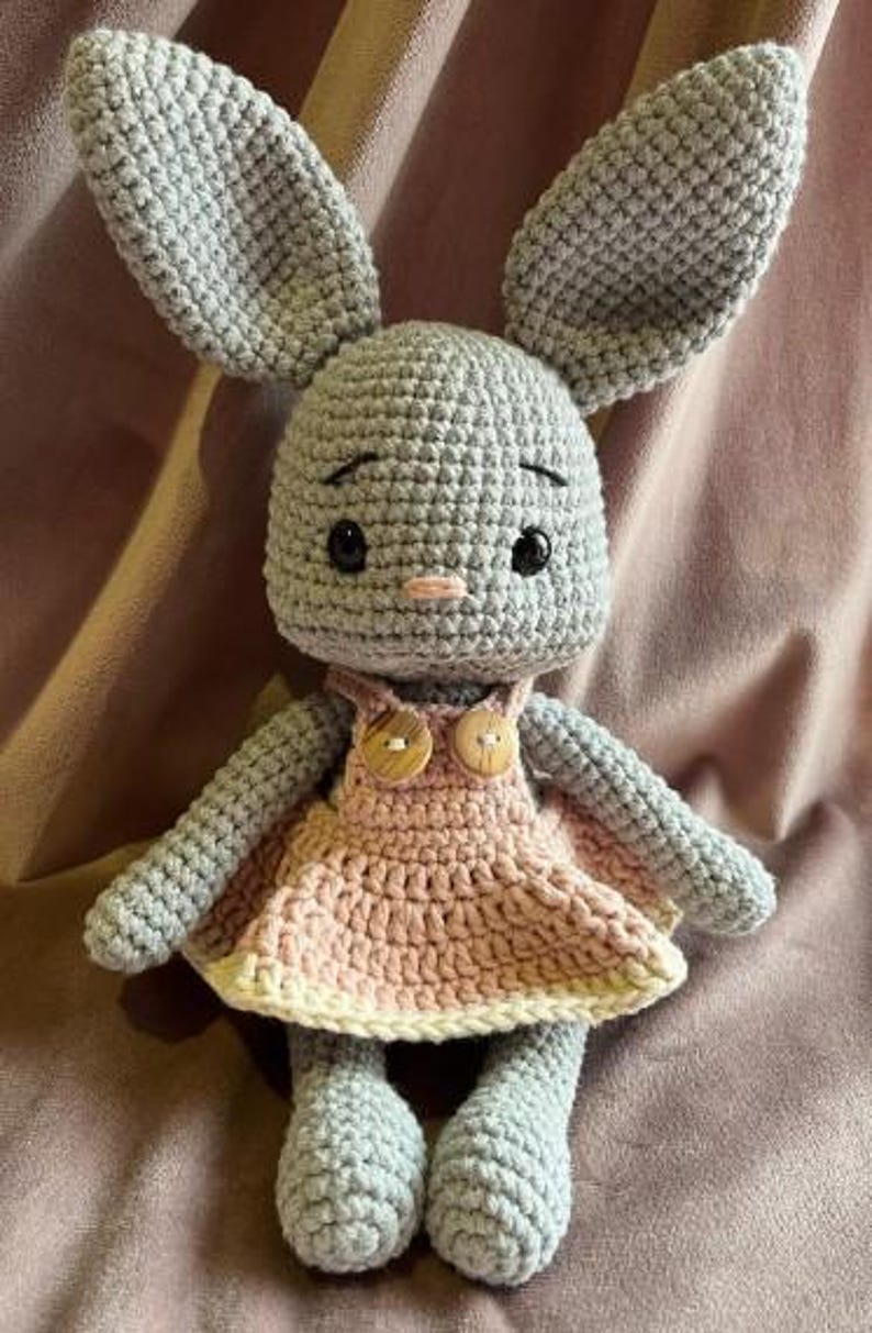 May include: A gray crocheted rabbit doll with long ears, black safety eyes, and a small pink stitched nose. The rabbit wears a light pink crocheted dress with a cream-colored hem and two round wooden buttons attached to the front straps.