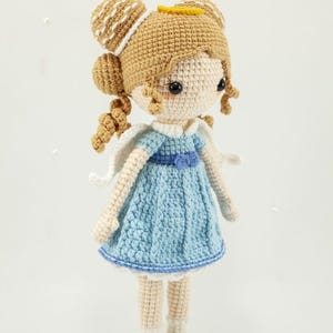 May include: A handcrafted crochet doll featuring light tan skin, blonde hair styled in two buns with small curls, and a blue dress with a white collar and wings. The doll wears white socks and yellow shoes, standing on a small circular base.