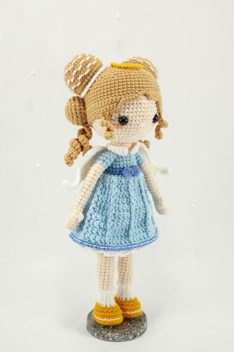 May include: A handcrafted crochet doll featuring light tan skin, blonde hair styled in two buns with small curls, and a blue dress with a white collar and wings. The doll wears white socks and yellow shoes, standing on a small circular base.