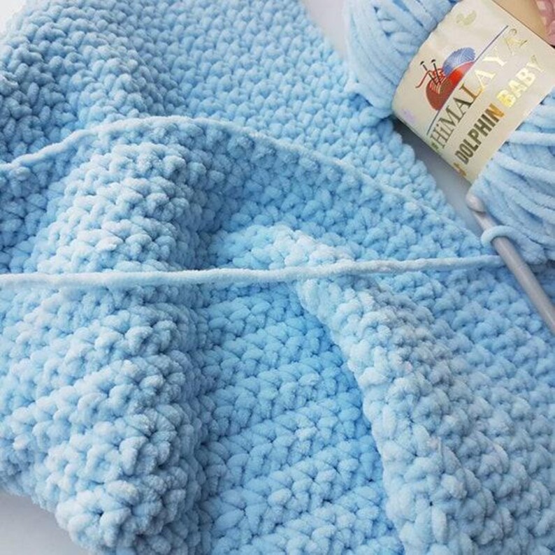May include: A close-up of a light blue, plush, crocheted blanket with a ball of yarn and a crochet hook. The yarn label reads "Himalaya Dolphin Baby". The blanket has a textured, soft appearance, suggesting a cozy handmade item.