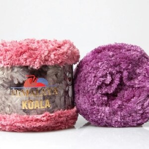 May include: Two skeins of Himalaya Koala yarn. One skein is wrapped in a paper label featuring a koala illustration and the text Himalaya Koala, with pink fuzzy yarn at the top and bottom. The second skein is a solid, textured purple yarn ball.