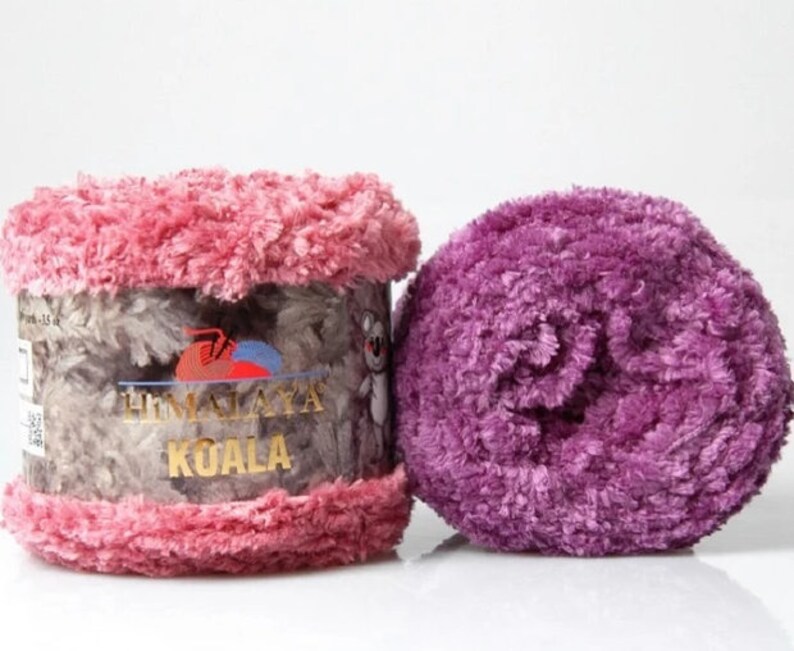 May include: Two skeins of Himalaya Koala yarn. One skein is wrapped in a paper label featuring a koala illustration and the text Himalaya Koala, with pink fuzzy yarn at the top and bottom. The second skein is a solid, textured purple yarn ball.