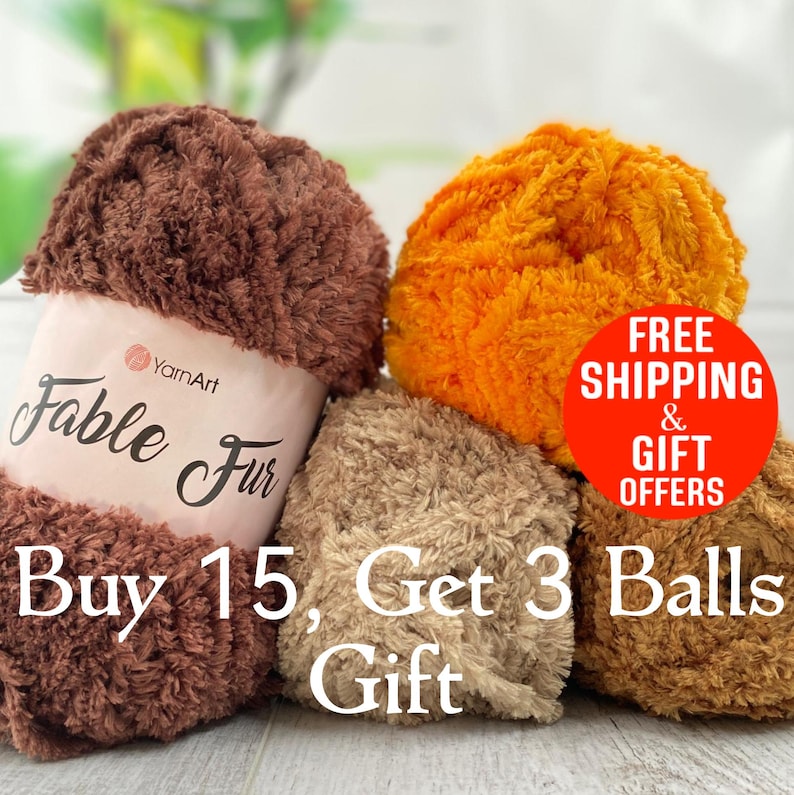 May include: Four balls of YarnArt Fable Fur yarn in brown, orange, and beige. The brown ball has a label reading YarnArt Fable Fur. Text on the image states: FREE SHIPPING & GIFT OFFERS, Buy 15, Get 3 Balls Gift. The yarn has a fuzzy texture.