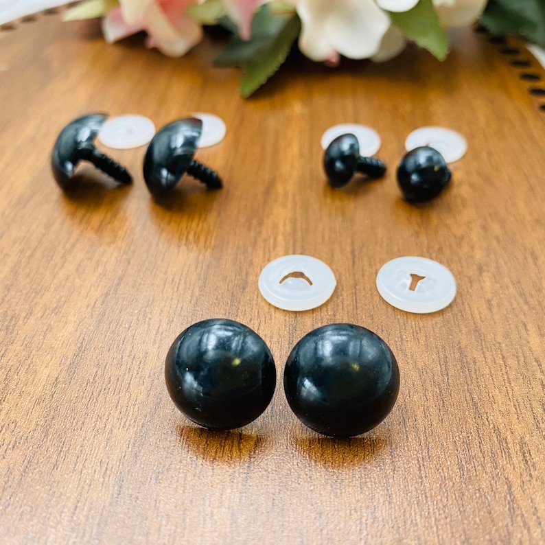 May include: A collection of black plastic safety eyes for stuffed animals and dolls. The set includes several smooth, round, black eyes with threaded posts and matching translucent white plastic washers to secure them in place.
