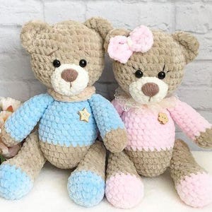 May include: Two handmade crochet teddy bears. One bear wears a blue sweater with a gold star, the other wears a pink dress and bow. Both have brown fur, white muzzles, and black eyes. The bears are approximately 12 inches tall.