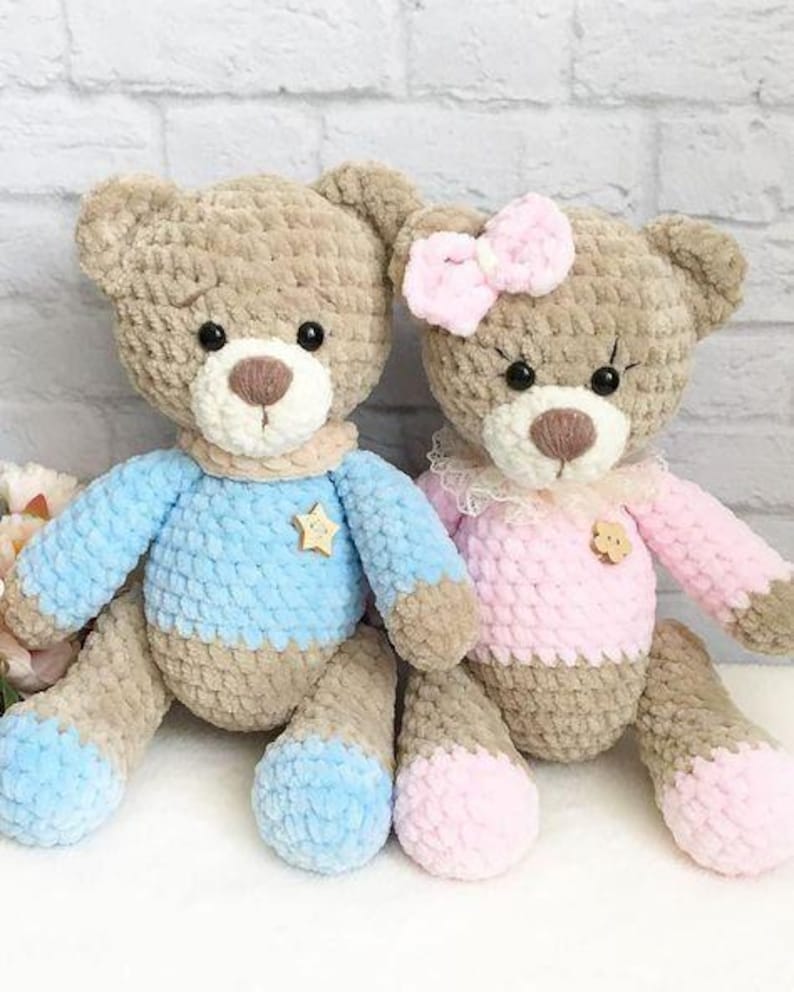 May include: Two handmade crochet teddy bears. One bear wears a blue sweater with a gold star, the other wears a pink dress and bow. Both have brown fur, white muzzles, and black eyes. The bears are approximately 12 inches tall.