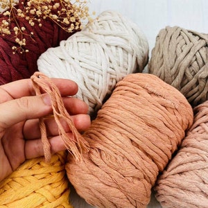 Pu&ograve; includere: Una collezione di bobine di corda di cotone intrecciato spesso in vari colori, tra cui bordeaux, bianco sporco, beige, pesca, giallo e rosa chiaro. Una mano tiene diversi fili sciolti della corda color pesca, mostrando la consistenza della fibra ritorta.