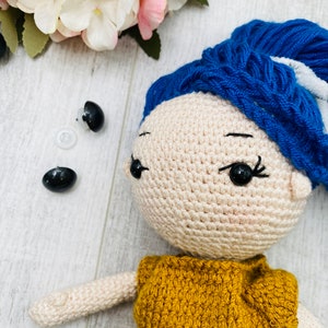 May include: A crocheted doll with beige skin, black plastic safety eyes, and vibrant blue yarn hair styled in a high bun. The doll wears a mustard yellow crocheted top. Two additional black plastic safety eyes and a clear washer are visible.