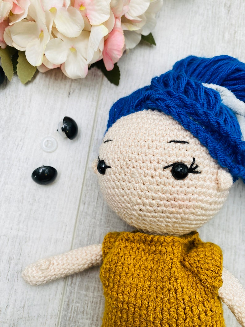 May include: A crocheted doll with beige skin, black plastic safety eyes, and vibrant blue yarn hair styled in a high bun. The doll wears a mustard yellow crocheted top. Two additional black plastic safety eyes and a clear washer are visible.
