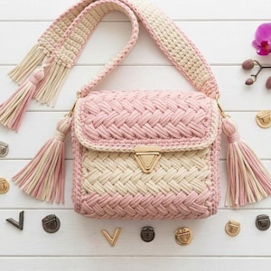 May include: A handmade crochet shoulder bag in pink and cream with a woven pattern. The bag features a flap closure with a gold-tone clasp, a matching strap, and decorative tassels. The bag is surrounded by decorative metal hardware and a pink orchid.