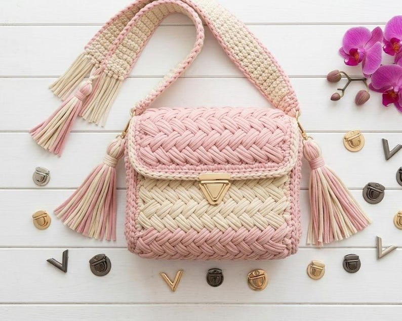 May include: A handmade crochet shoulder bag in pink and cream with a woven pattern. The bag features a flap closure with a gold-tone clasp, a matching strap, and decorative tassels. The bag is surrounded by decorative metal hardware and a pink orchid.