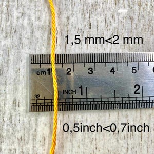 May include: A single strand of bright yellow twisted cord positioned vertically against a metal ruler. The text 1.5 mm < 2 mm and 0.5 inch < 0.7 inch is printed on the image, indicating the thickness of the cord relative to the measurement tool.