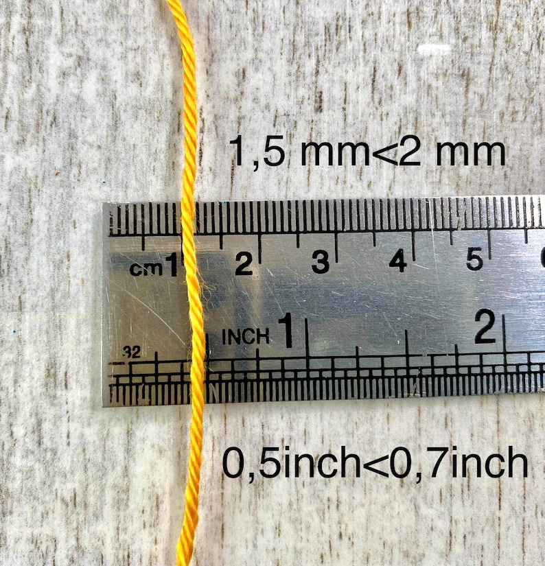 May include: A single strand of bright yellow twisted cord positioned vertically against a metal ruler. The text 1.5 mm < 2 mm and 0.5 inch < 0.7 inch is printed on the image, indicating the thickness of the cord relative to the measurement tool.