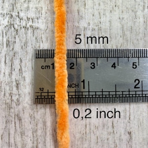 May include: Close-up of a vibrant orange chenille stem, approximately 0.2 inches in diameter, positioned next to a ruler. The ruler displays both inch and centimeter measurements. The text "5 mm" and "0,2 inch" are visible.