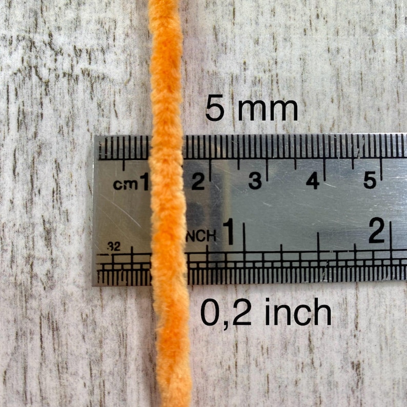 May include: Close-up of a vibrant orange chenille stem, approximately 0.2 inches in diameter, positioned next to a ruler. The ruler displays both inch and centimeter measurements. The text "5 mm" and "0,2 inch" are visible.