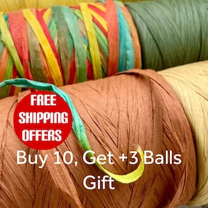 May include: Multiple spools of raffia ribbon in various colors including solid olive green, solid tan, and a multi-colored spool featuring alternating strands of red, yellow, teal, and brown. The texture is fibrous and matte with a paper-like appearance.