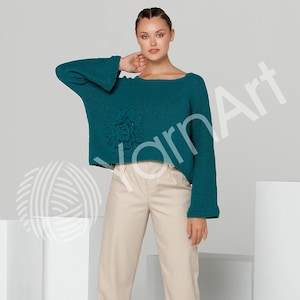May include: Teal-colored sweater with a floral applique. The sweater has long sleeves with flared cuffs and a wide neckline. The model is wearing beige pants. The background is a light gray.
