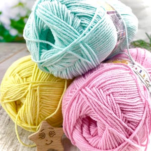 May include: Three skeins of soft yarn in pastel mint green, bright yellow, and light pink. Each skein features a paper label with printed text and graphics. A small, light-colored wooden star ornament with a carved smiling face is tucked in front.