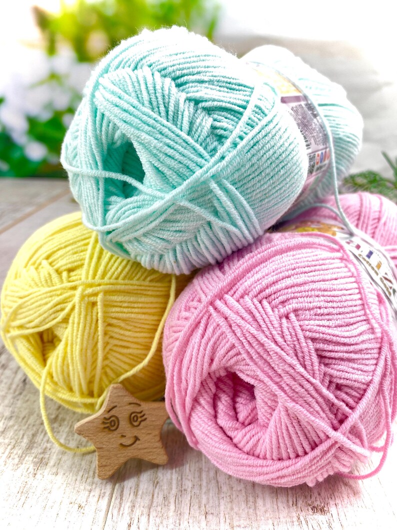 May include: Three skeins of soft yarn in pastel mint green, bright yellow, and light pink. Each skein features a paper label with printed text and graphics. A small, light-colored wooden star ornament with a carved smiling face is tucked in front.