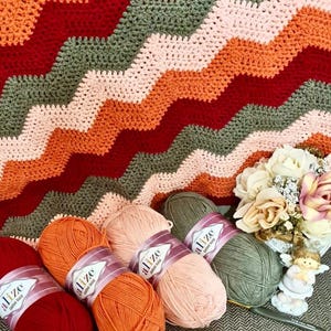 May include: A crocheted blanket featuring a chevron pattern in alternating stripes of dark red, orange, light pink, and olive green. Beside the blanket are four skeins of Alize Cotton Gold yarn in matching colors and a small decorative angel figurine.