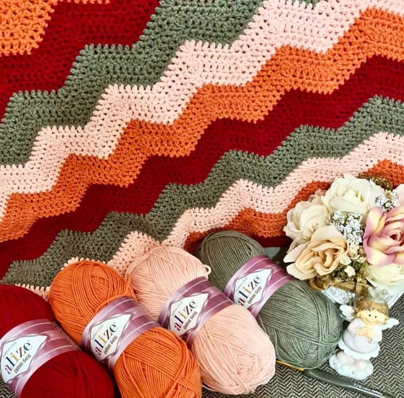 May include: A crocheted blanket featuring a chevron pattern in alternating stripes of dark red, orange, light pink, and olive green. Beside the blanket are four skeins of Alize Cotton Gold yarn in matching colors and a small decorative angel figurine.
