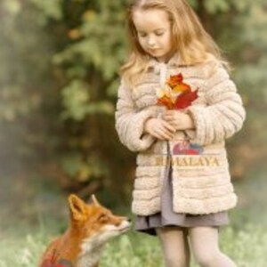 May include: A child with long blonde hair wears a beige textured jacket over a grey dress, holding a cluster of autumn leaves. A red fox stands on the grass nearby. The word HIMALAYA is printed in gold on the front of the jacket.