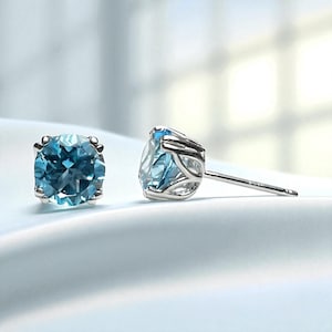May include: A pair of stud earrings featuring round, light blue gemstones set in a silver metal. The gemstones have a faceted appearance, and the silver setting has a decorative design around the stone. The earrings are displayed on a white surface.