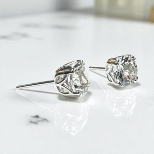 May include: A pair of silver stud earrings with large, clear, round-cut gemstones. The earrings have a detailed, openwork design around the gemstone setting. The posts are thin and silver. The earrings are displayed on a white surface.