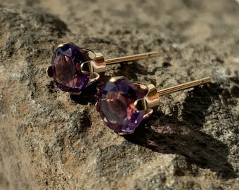 May include: A pair of gold stud earrings with round, faceted, purple gemstones. The earrings are set on a textured, gray-brown stone surface, with the sunlight reflecting off the gemstones and gold posts.